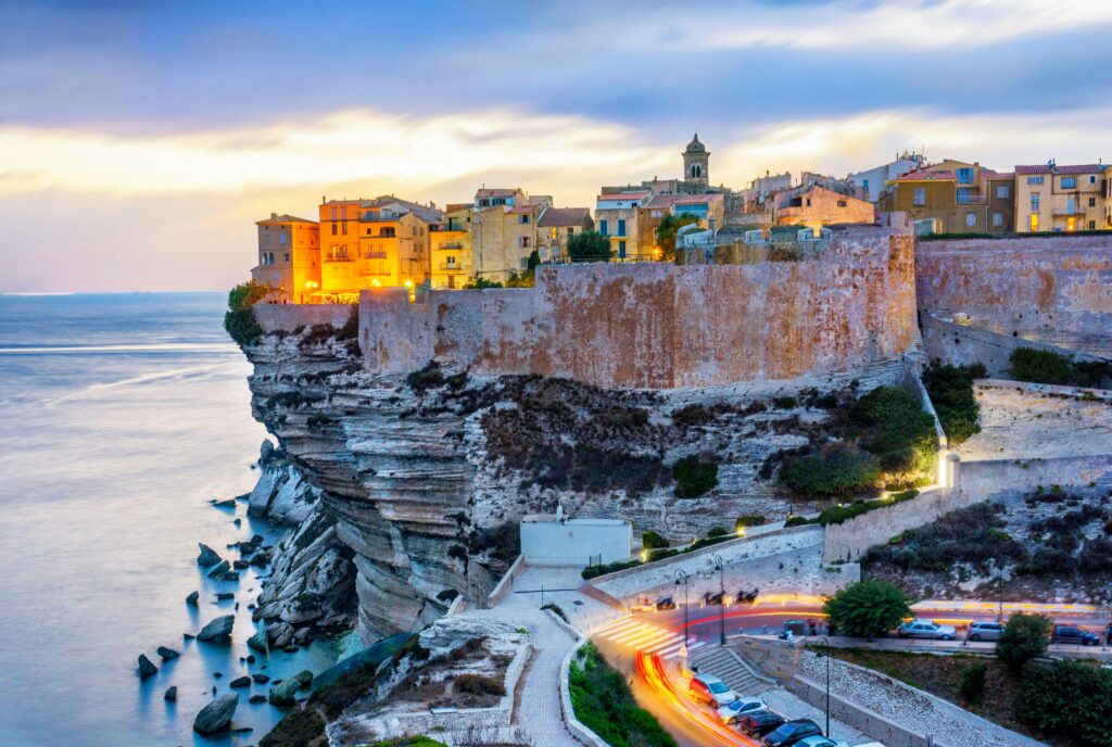 A luxury catamaran sailing past the historic limestone cliffs and Genoese citadel of Bonifacio, Corsica, at sunset.