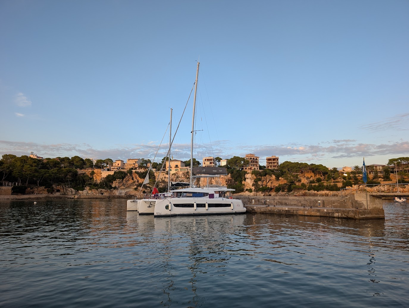 A modern luxury catamaran docked at a stone pier in a calm Mediterranean bay with coastal villas in the background at sunset.