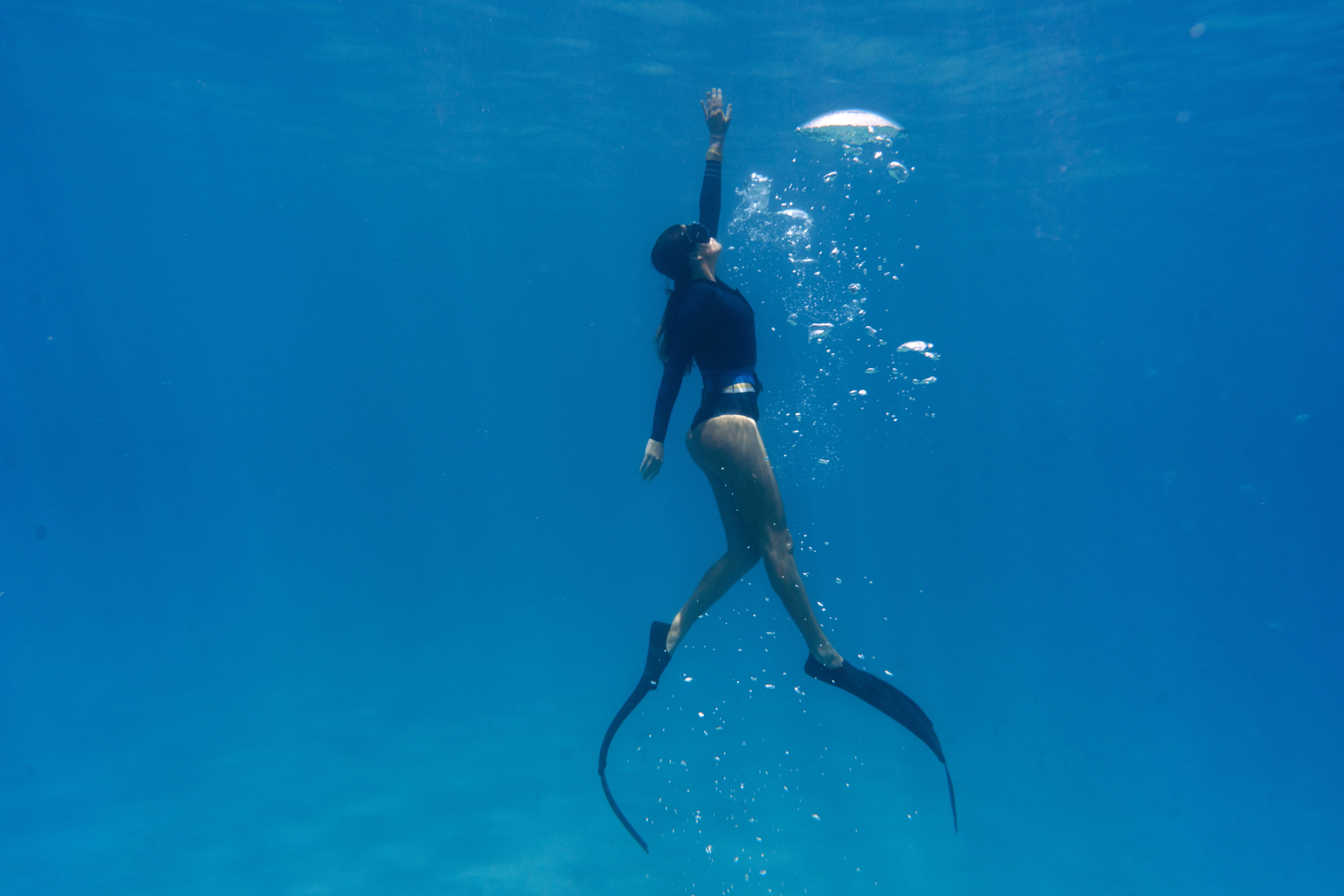 A female freediver in a black wetsuit using long carbon fins to ascend toward the surface in turquoise waters.